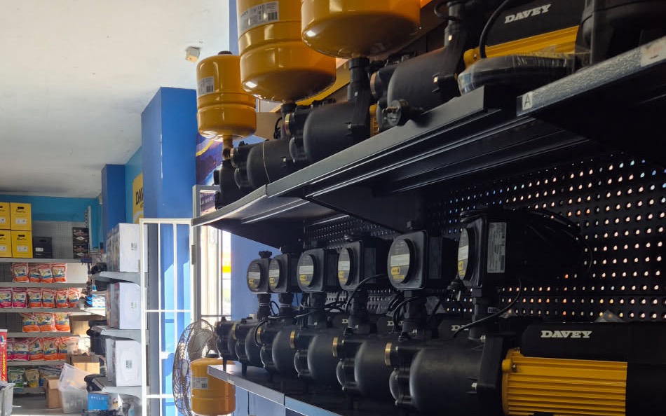 Inside the shop, the pump wall: rows of Davey pressure pumps racked on steel shelving with yellow pressure tanks mounted above them, and the front door open to the street beyond.