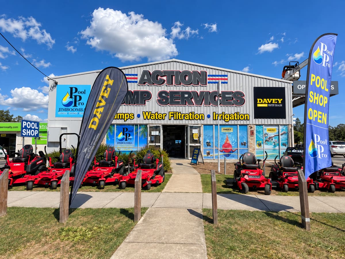 The Action Pump Services shopfront on Tamborine Street in daylight, its signage reading pumps, water filtration and irrigation, with the Pool Pro Jimboomba sign alongside, a Davey water dealer sign, Davey and Pool Pro flags either side of the path, and a row of Gravely ride-on mowers out the front.